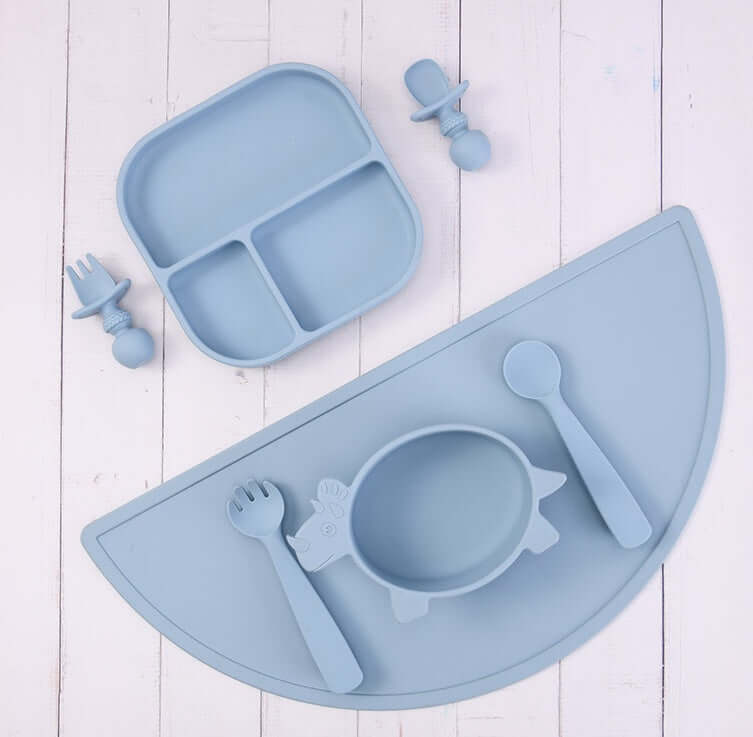 Silicone baby tableware set with placemat, sectioned plate, bowl, knife, fork, and baby food accessory tools in pastel blue color.