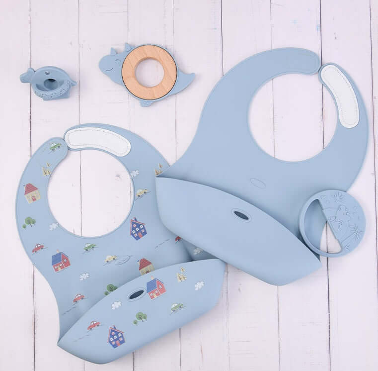 Silicone baby food accessory tools including bibs, teething ring, and a bird-shaped toy for infants over 3 months old on a white wooden background