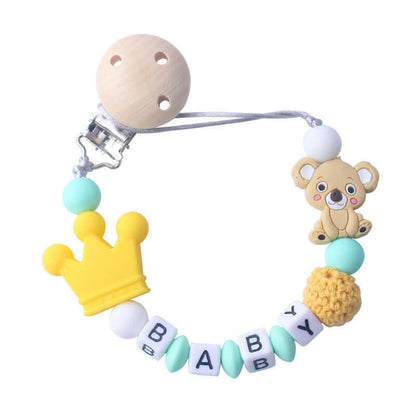 Baby pacifier clip with wooden bead, silicone koala bear, yellow crown, and "BABY" letter blocks on white background.
