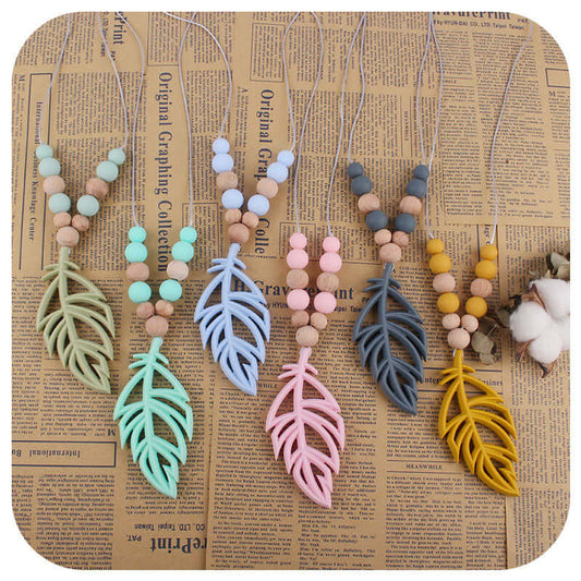Silicone leaf teether necklaces in mint green, bean green, pink, gray, turmeric, and blue displayed on vintage newspaper background.