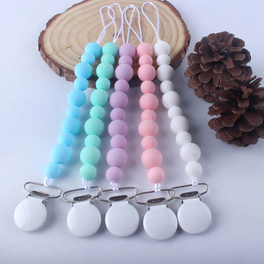 Silicone pacifier clips in pink, blue, green, purple, and white, laid on a wooden surface next to pine cones, ideal for babies over 3 months.