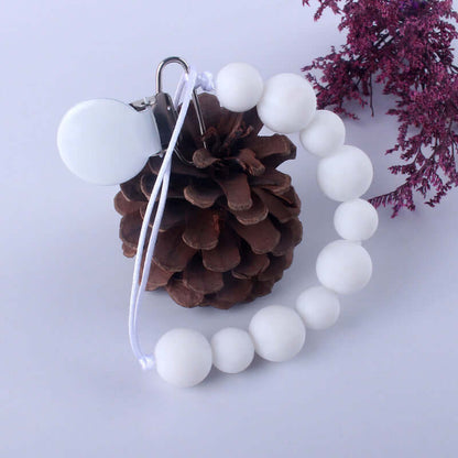 White silicone pacifier clip with round beads, ideal baby product to prevent pacifier loss, displayed with pinecone and purple flowers.