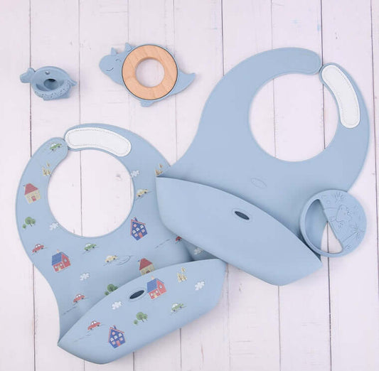 Silicone baby food accessory tools including bibs, teething ring, and a bird-shaped toy for infants over 3 months old on a white wooden background