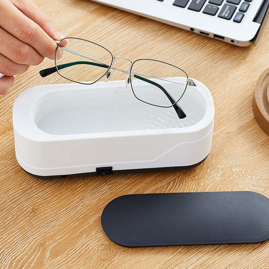 Person using a white multifunctional ultrasonic eyeglasses cleaner on a wooden desk with removable lid nearby.